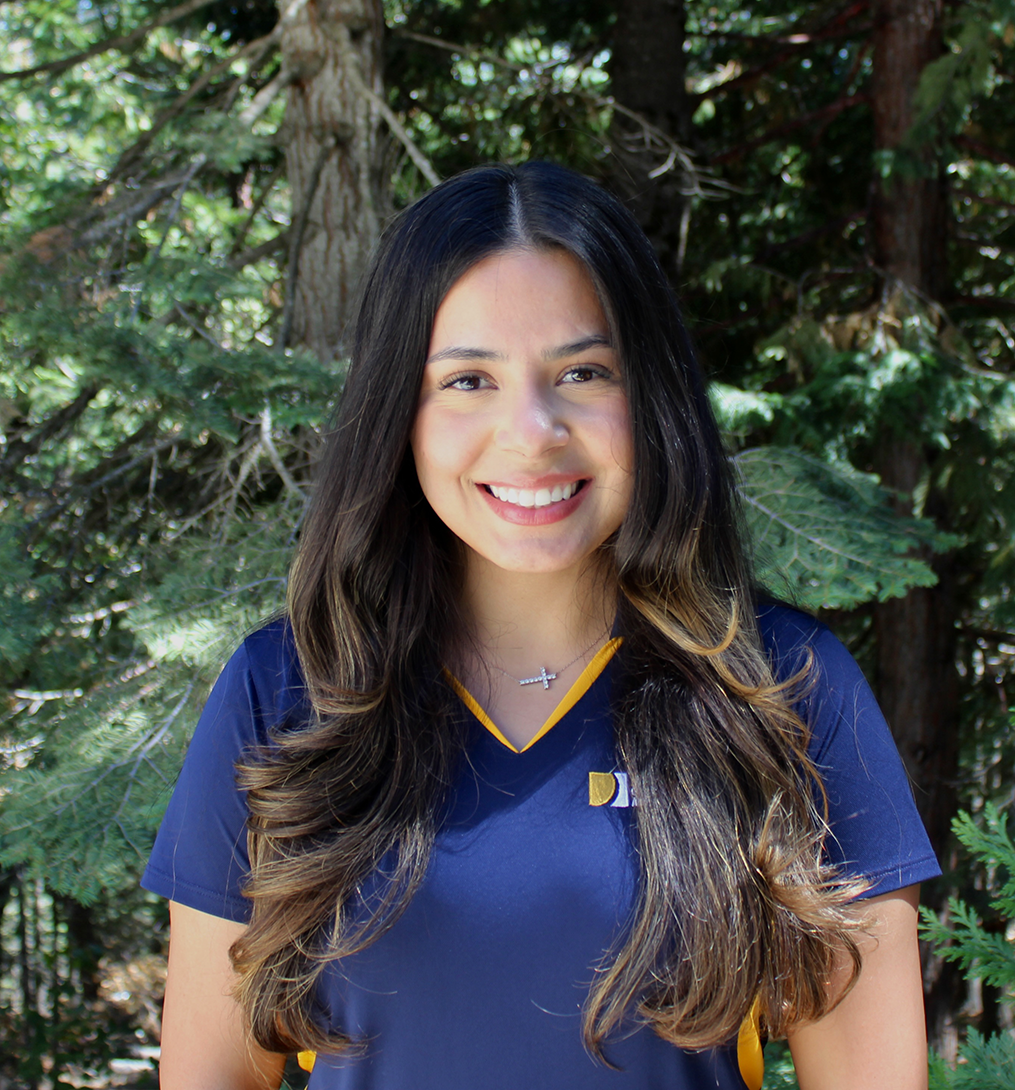 Smiling young woman with long dark hair in navy shirt, evergreen trees behind her