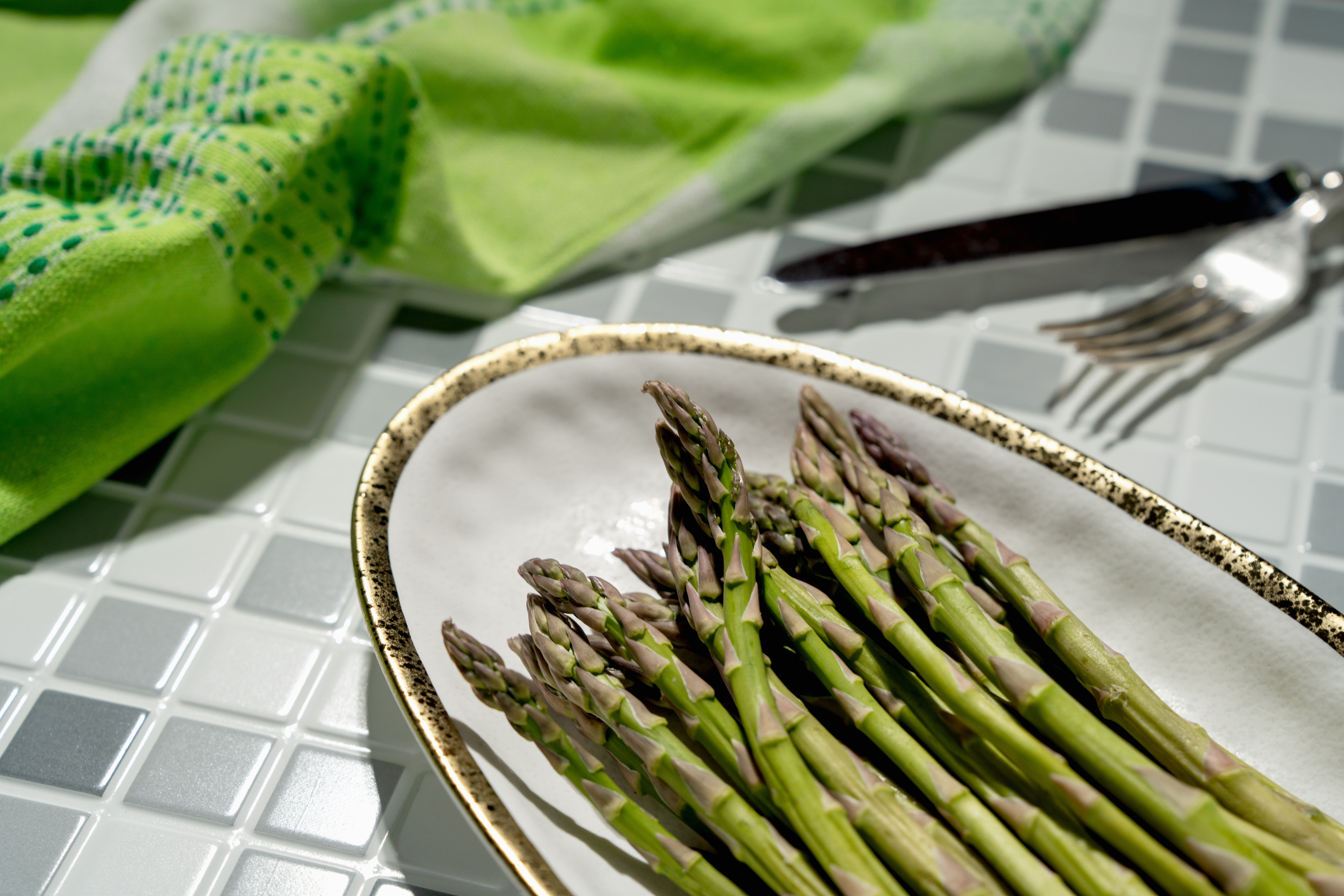 Fresh asparagus on a plate with a fork and knife in the background next to a cloth napkin.