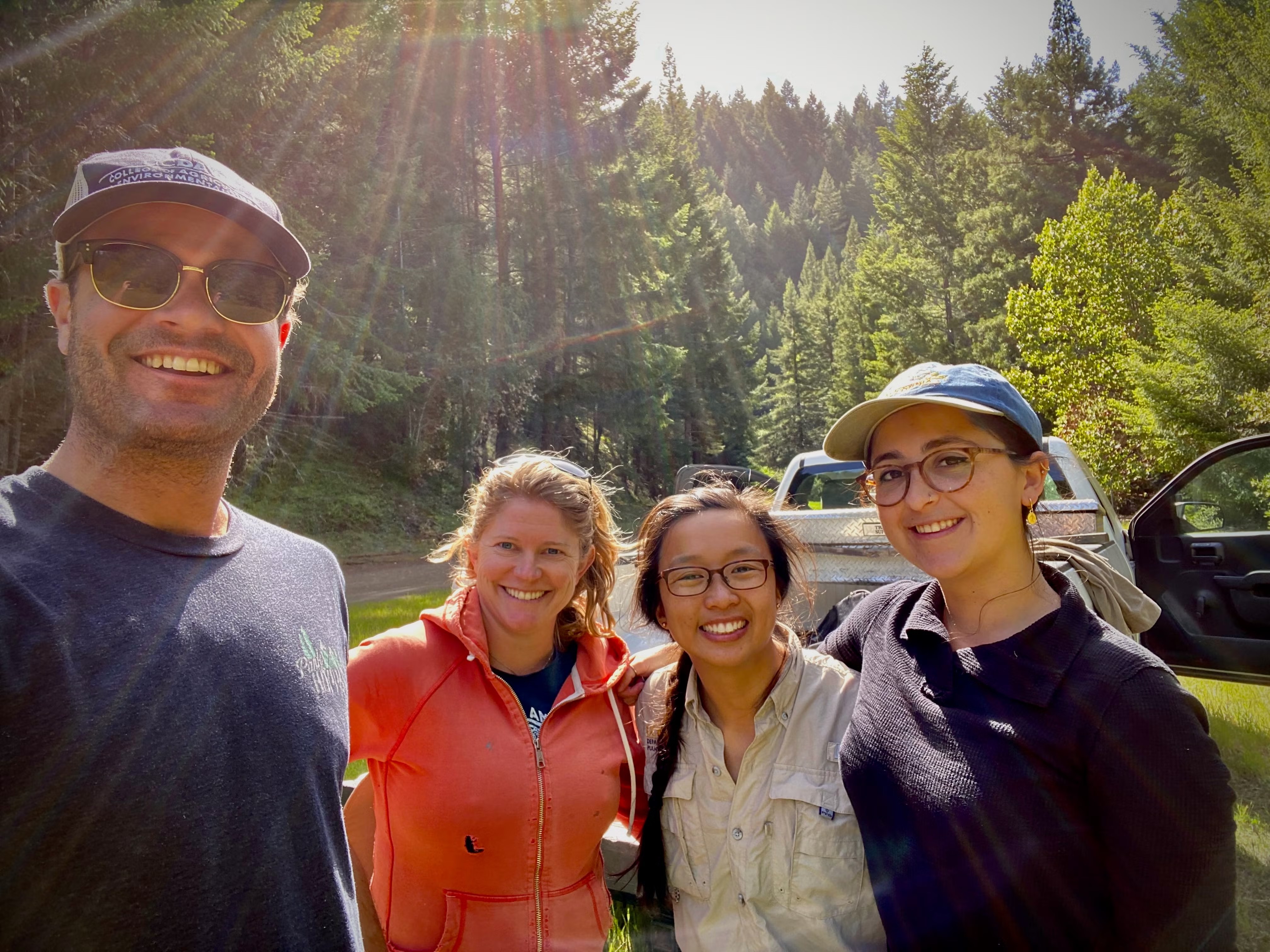 Four smiling people taking a sunny forest selfie with lens flare and a parked car