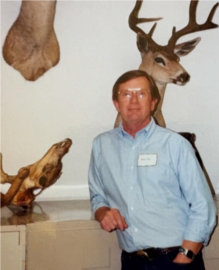 A person with a name tag leans on a table with mounted animals in the background and beside him.