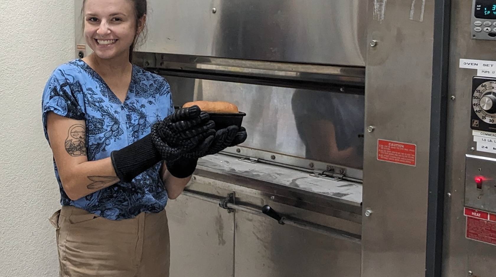 A woman holds a loaf of fresh baked bread in the tin beside an industrial oven.