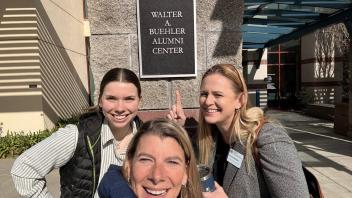 Three smiling women pose for a selfie in front of a building sign.