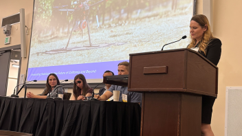 Several people sit at a table behind microphones while another person stands behind a podium.