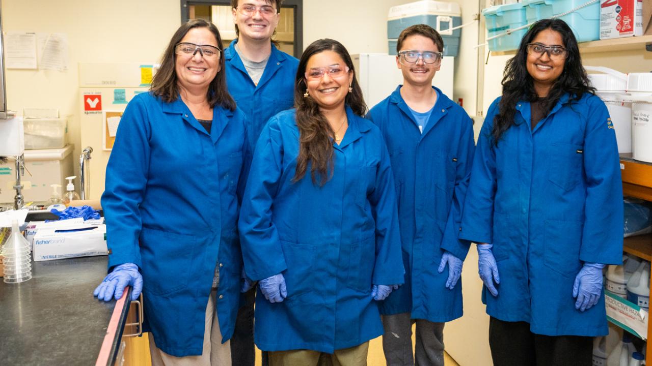 Five researchers wearing blue lab coats and gloves, smiling in a busy laboratory