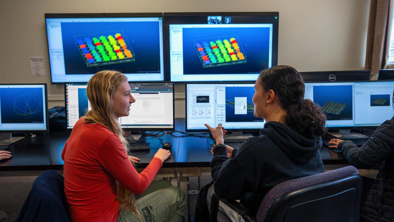Two women engage in conversation at desks with multiple computer monitors displaying colorful graphics.