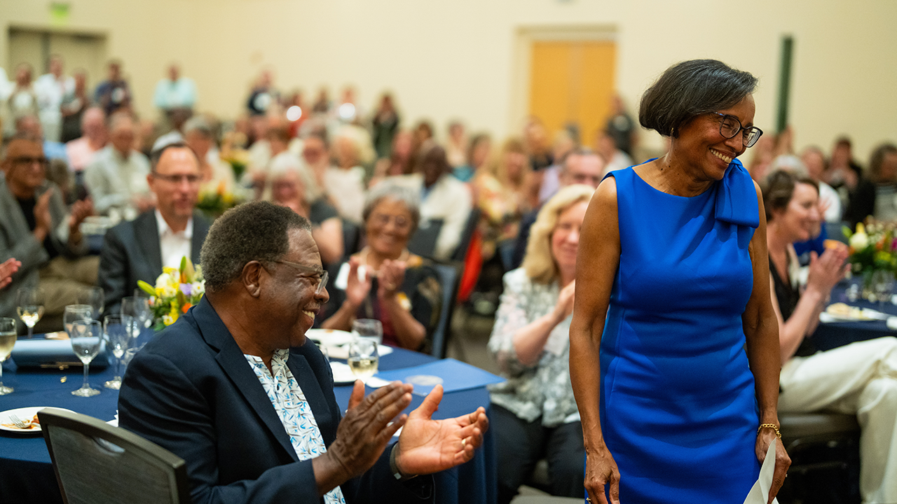 A woman stands and smiles as people around her applaud her.