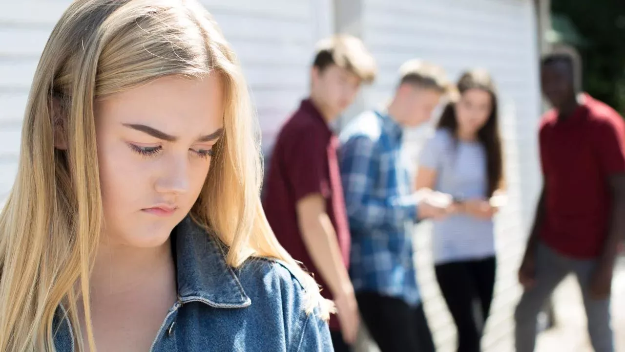 A teenage girl looking downward while standing apart of from a crowd of teens.