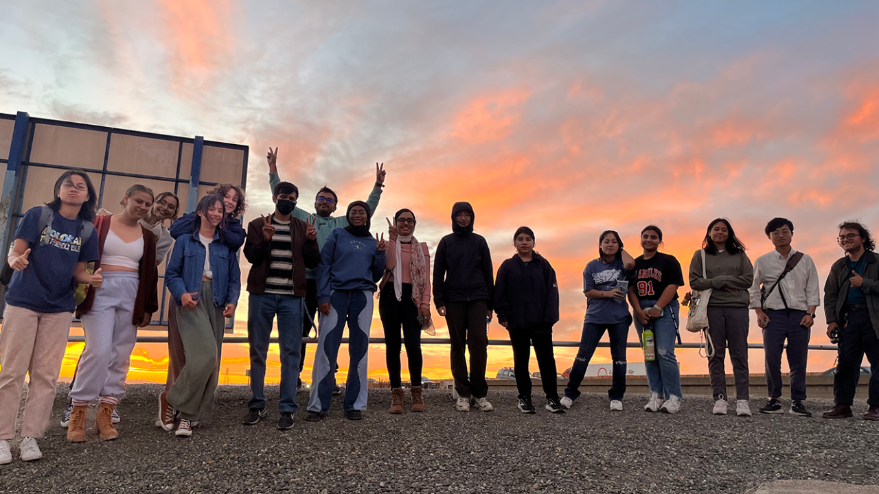 The image shows a group of people standing together, silhouetted against a stunning sunset or sunrise.
