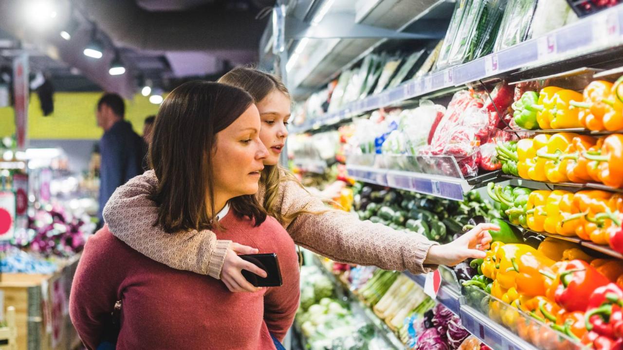 A woman, holds a child on her back, while they look together at vegetables in the produce section of a grocery store.