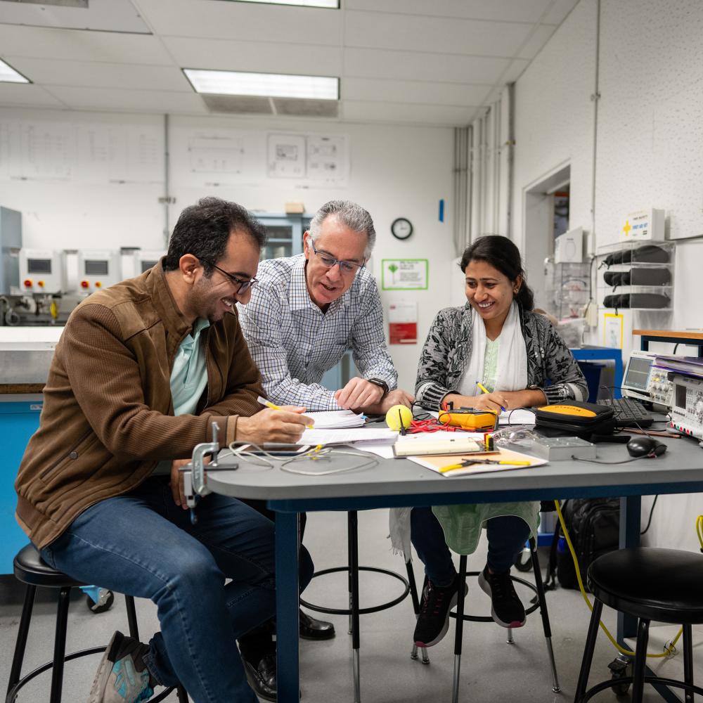 Three people sit at a desk and look over machinery and paperwork together.