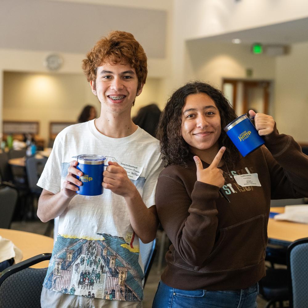 Two students stand back to back and hold up their mugs from Aggie Jumpstart.