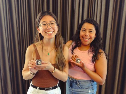 Two people smiling and holding pins, standing against a brown curtain backdrop.