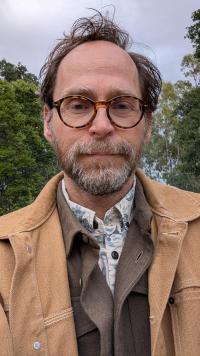 Outdoor headshot of man with glasses and beard, tan jacket, trees behind