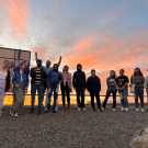 The image shows a group of people standing together, silhouetted against a stunning sunset or sunrise.