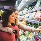 A woman, holds a child on her back, while they look together at vegetables in the produce section of a grocery store.