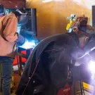 Bright sparks are visible from the welding process, indicating that metal work is being performed. The individuals are wearing protective gear, including helmets with visors and gloves, which is essential for safety during welding.