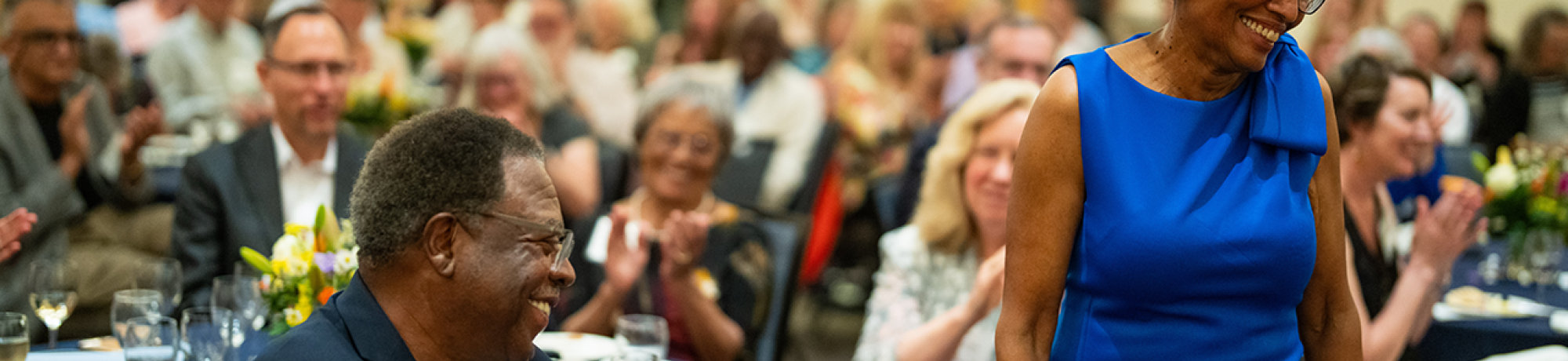 A woman stands and smiles as people around her applaud her.