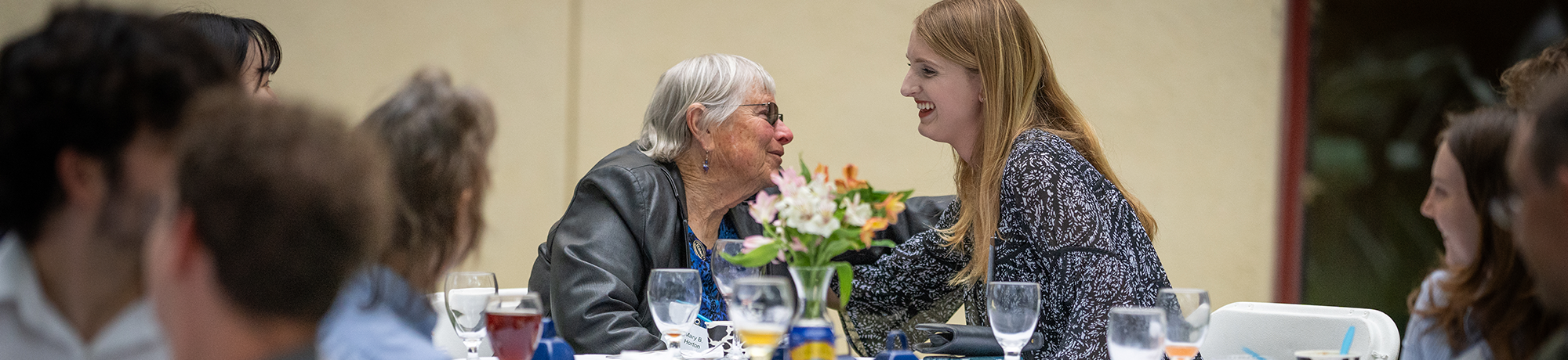 A student hugs a woman while seated at a dining table.