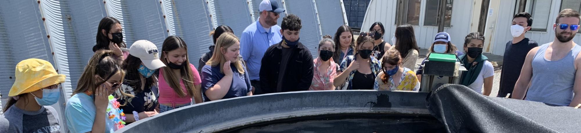 Masked group gathered around large outdoor circular water tank beside a greenhouse